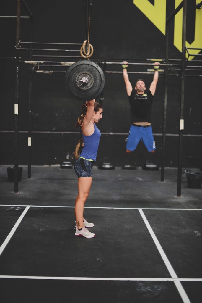 A woman lifting weights and a man doing pullups in a CrossFit gym