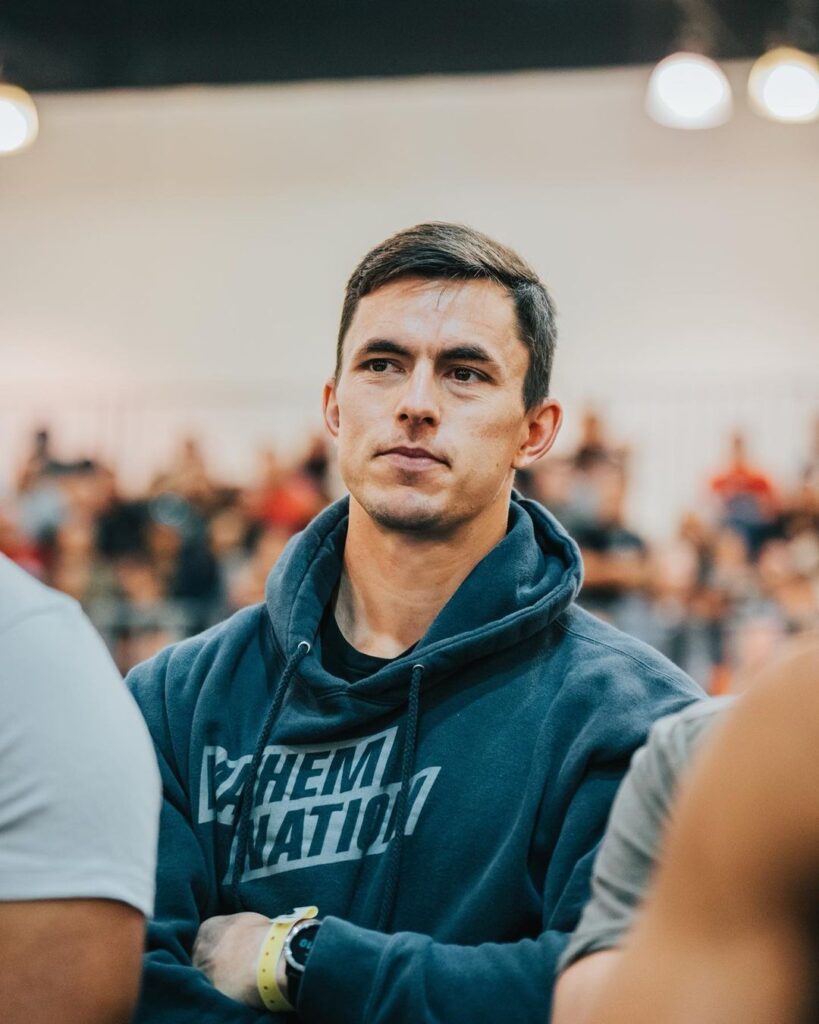 Jake Lockert, Director of Online Programming for Mayhem Athlete, wearing a hoodie and attentively observing a CrossFit event.
