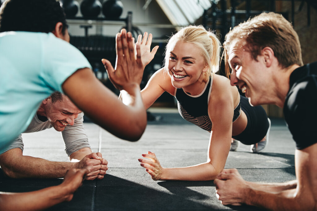 Two gym members out of a group of friends high fiving while planking on a gym floor.
