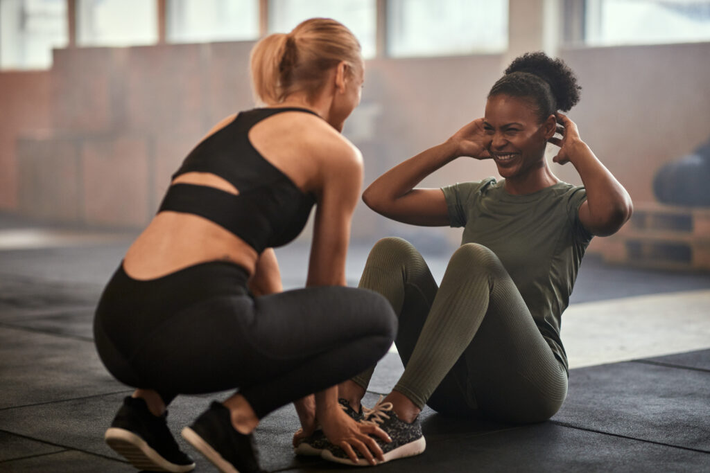 A female trainer helping a young woman while doing sit-ups in a gym.