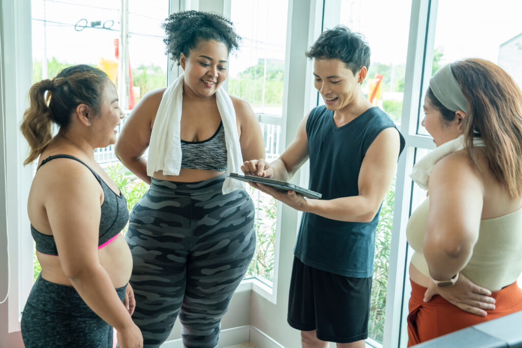 Three female gym members in workout attire standing together in a bright gym space, smiling and looking at a tablet as the trainer explains their performance data on the screen.