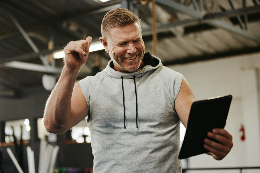 A coach in a gym smiles and raises his fist in celebration while looking at a tablet, reacting to positive fitness coaching and performance results.