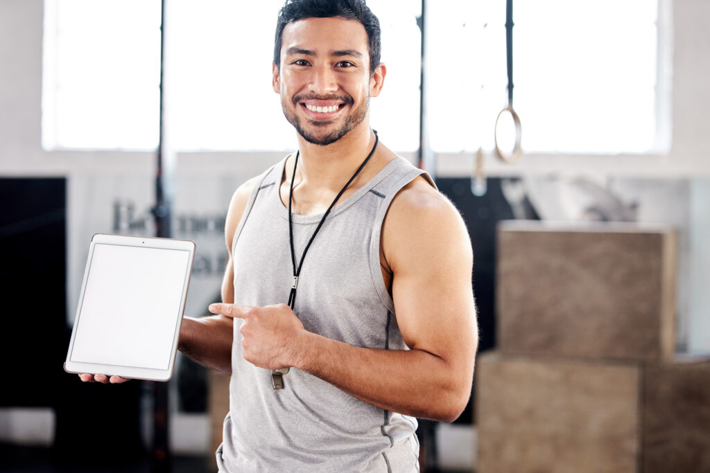 Fitness trainer in a gym smiles while holding and pointing to a tablet and presenting fitness coaching plans on gym management software.