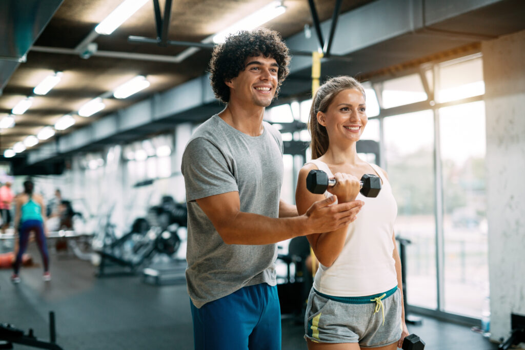 Personal trainer coaches a woman on consistency through a dumbbell curl, guiding her form during a workout in a bright gym.